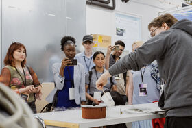 Group watches a lab demo as a person in grey hoodie pours from cup into steaming pot on table