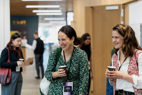 People with badges chat over coffee in a bright conference hallway, others networking in the background