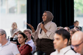 Person in brown headscarf speaks into a microphone while standing among a seated audience indoors