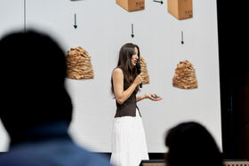 Woman in white skirt presenting on stage, gesturing to slides with stacked cardboard and arrows