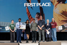 Group on stage with a FIRST PLACE banner; one person holds a diploma-sized certificate