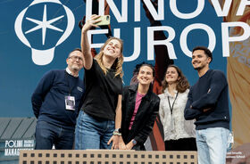 Group of young adults take a selfie on stage at an innovation event, standing before a large INNOVA EUROPE screen.