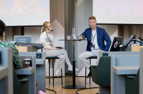 Two speakers sit at a tall round table in a lecture hall, talking with headsets and laptops nearby.