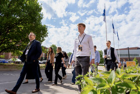 Group of conference visitors walking outside by trees, buildings and flagpoles under a cloudy sky