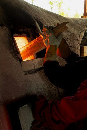 Person in gloves feeding firewood into a glowing kiln with orange light