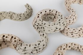 Curving rings of textured beige and grey clay with perforated, wavy surfaces on a white background.