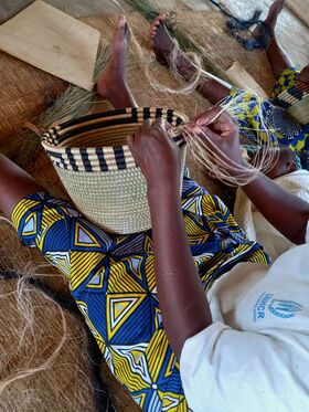 Person weaving a striped basket by hand, seated on a mat, wearing patterned blue and yellow skirt