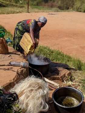 Woman in patterned dress pours water from yellow jerrycan into steaming pot outdoors on dusty ground
