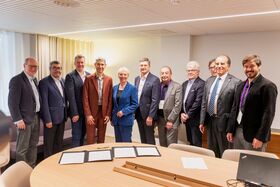 Ten suited adults stand smiling in a meeting room, with documents laid out on a round wooden table.