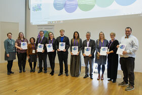 Ten adults in formal clothes stand indoors on wooden floor, several holding white award certificates.