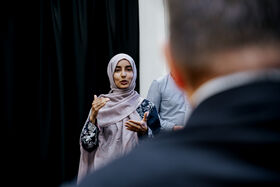 Woman in a light pink hijab speaking to a group indoors, gesturing with her hands