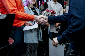 People at an award event shaking hands, holding certificates and red roses in a crowded indoor setting