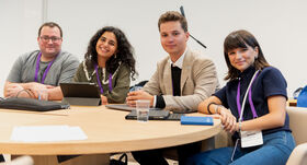 Four young adults sit at a round meeting table with laptops, tablets and notes, smiling at the camera.