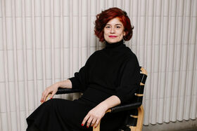 Lyhyttukkainen punatukkainen henkilö mustissa vaatteissa istuu tuolilla vaaleaa uritettua seinää vasten