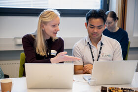Two colleagues discuss work at laptops in a bright office, one gesturing while the other listens.