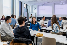 People in a bright classroom sit in small groups at white tables, talking during a workshop.