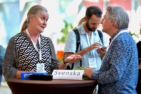 Two people talking at a round table marked “Svenska”, others with badges and phones in the background