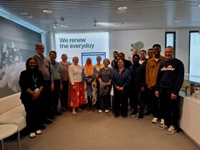 Diverse group of people smiling in an office, standing under a sign reading “We renew the everyday”.