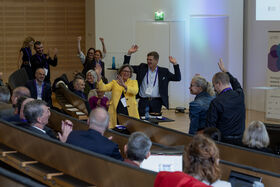 Conference audience in a lecture hall laughing and raising hands; woman in yellow jacket stands in the centre