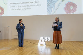 Two women with microphones on a wooden stage in front of a projected award slide and glowing letter A sculpture.