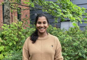 Piushana Abeygunawardena ruskeassa neuleessa seisoo vihreiden pensaiden ja talon edessä