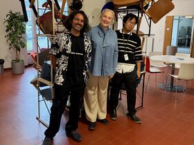 Three people smiling in a casual office lounge, standing before a metal rack stacked with chairs.