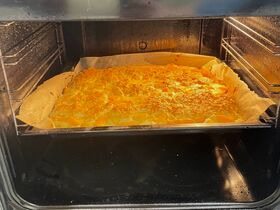 Rectangular golden traybake with cheese and sliced vegetables baking on parchment in an oven