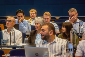 Mixed group of adults in blue lecture hall seats, listening and talking during a seminar with laptops and bottles