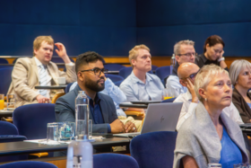 Adults in a blue lecture theatre sit at desks with laptops and drinks, listening attentively to a talk