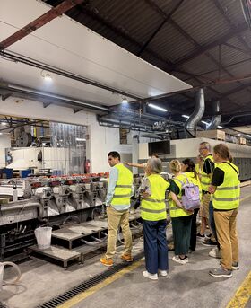 Group in yellow safety vests touring an industrial factory, watching a large silver machine in operation