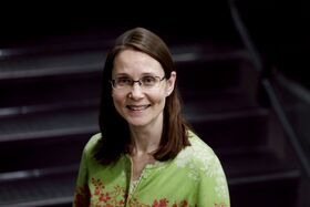 Smiling Sari Kujala with glasses and long brown hair, in green patterned top, standing indoors on dark stairs