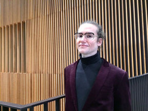 silmälasipäinen mies seisoo puurimoista tehdyn seinän edessä yllään violetti pikkutakki