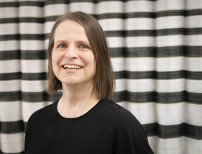 mustapaitainen nainen hymyilee, taustalla raidallinen verho