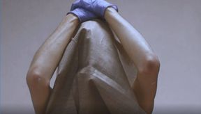 Henkilö tiukasti ylävartalon rajaavassa kuvassa. Henkilö pitää käsiään päänsä päällä, päässään / vartalon peittona maalauskangas. Kädessä henkilöllä on sinertävät kumihanskat, käsissä valkoisia maalitahroja. Kuvan tausta on harmaa.