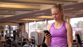 Nainen on kuntosalilla ja katsoo hymyillen puhelinta, jota pitää kädessään.