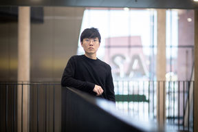 Vietnamista kotoisin oleva opiskelija on pukeutunut mustiin vaatteisiin ja nojaa kaiteeseen Tietotekniikan talolla. Taustalla näkyy valoa ja Aalto-yliopiston logo.