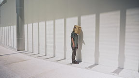 Still from the film Like Shadows Through Leaves, 2021. A figure stands next to a minimalist wall wearing a cardboard bird mask