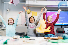 Kolme lasta kädet ilmassa luokkahuoneessa, värikkäiden paperitöiden ympäröimänä.