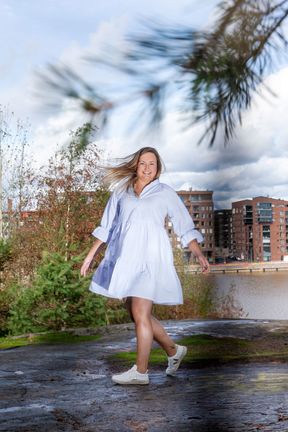 Laura Sivula valokuvattuna ulkona, kaupunkimaisemassa, taustalla näkyy kerrostaloja, kuvan oikeassa yläkulmassa on männyn oksa, Sivula on pukeutunut mekkoon, jonka helma hulmuaa, hänellä on jalassa valkoiset tennarit.