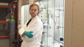 A person in a lab coat and green gloves stands in a science laboratory with various equipment and machinery.