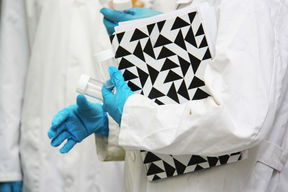 A person with a lab coat and protective gloves carrying test tubes and a black-and-white notebook. Their face is not pictured.
