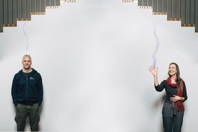 Jani Romanoff ja Maria Joutsenvirta seisovat hymyillen ja rinnakkain pienen välimatkan päässä toisistaan valkoisen seinän edessä. Kuvan yläosassa näkyy kuparipintainen portaikko. Joutsenvirta näyttää heiluttavan oikeaa kättään, josta lähtee kuvanmuokkauksella luotu sähkövirtaa muistuttava vaaleansininen viiva kiertäen yläkautta kohti Jani Romanoffin päätä. Kuva: Jaakko Kahilaniemi.