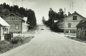 Suomalaisia taloja, neuvostoaikainen kirjakauppa, televisioantenneja ja sähkötolppia Kurkijoen taajaman pääkadun Ulitsa Leninan varrella. Pöllyävää hiekkatietä pitkin lähestyy vaalea henkilöauto.