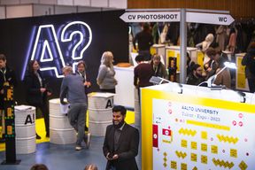 A lot of people, fair walls and signs in the picture. A large black wall with logo A? in the middle and people standing in front of it.