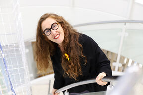 Hymyilevä silmälasipäinen nainen pitkät hiukset auki seisoo portaikossa ja katsoo ylöspäin kameraan