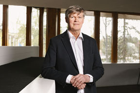 Tapani Vuorinen in a black suit, facing camera in front of large windows.