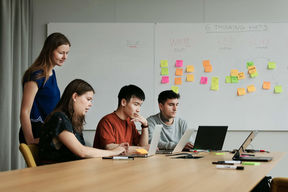 A teacher standing behind three students working on their laptops. 