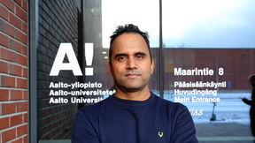 Ashish Thapliyal in a dark blue shirt stands in front of an Aalto University entrance with brick wall and glass door.