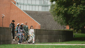 A group of people wearing colourful student overalls walk down steps beside a brick building and a large tree.