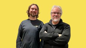 Two people standing against a yellow background; one in a dark shirt, the other in a dark jacket with arms crossed.